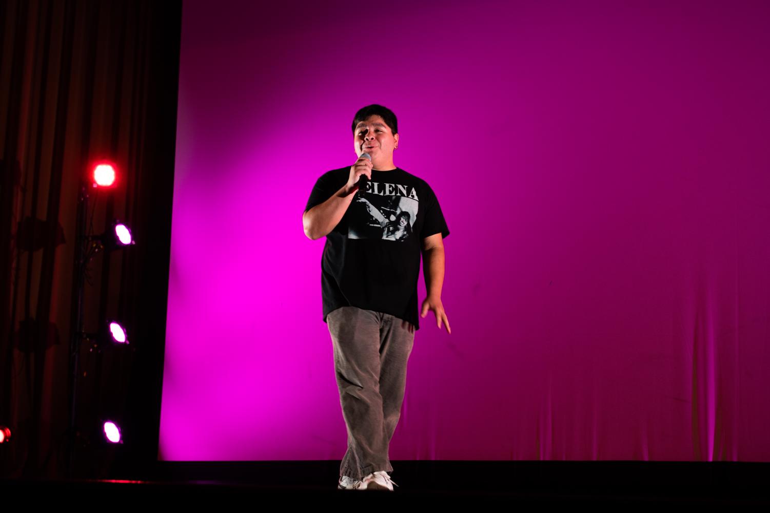 An emcee wearing a dark Selena shirt holds a microphone onstage. 