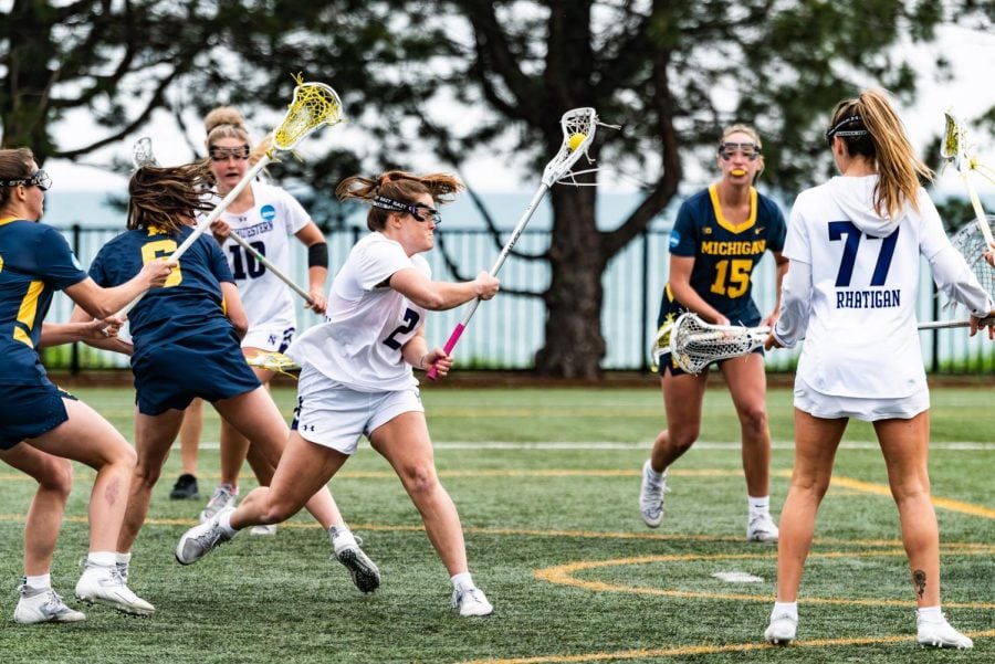 A lacrosse player cradles the ball in her stick and turns her back to her defenders.