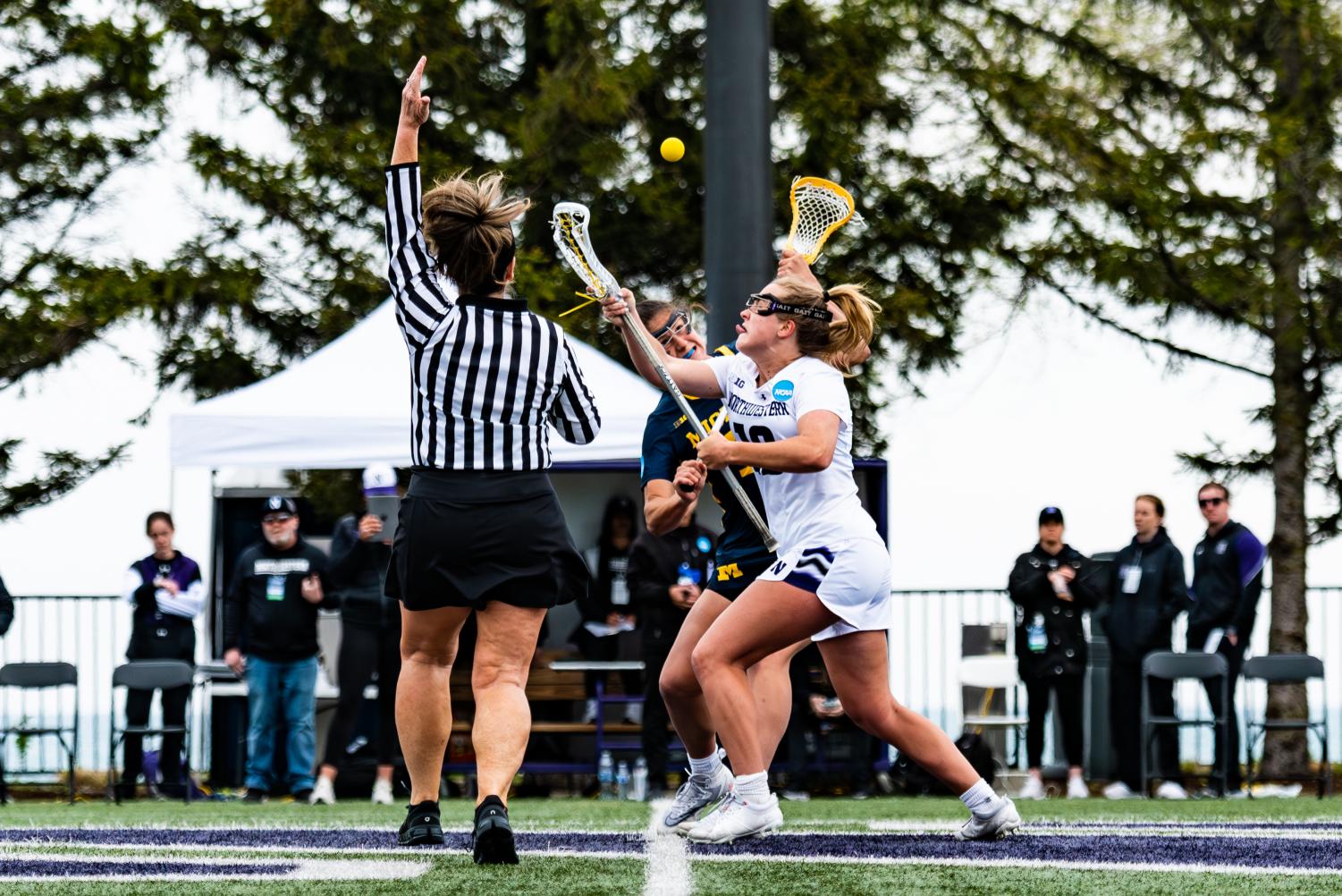 Two lacrosse players raise their sticks toward a ball as a referee points in the air.