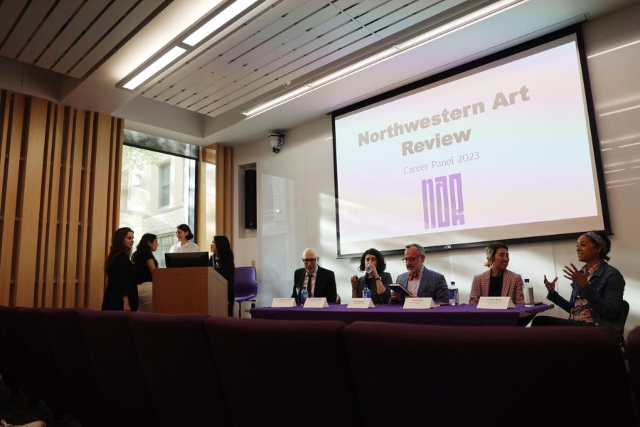 Four people stand around a podium on the left side of the photo. Five people sit behind a long table, with a screen reading Northwestern Art Review Career Panel 2023 behind them.