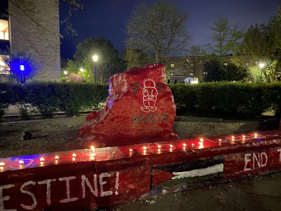 A night-time photograph of a large boulder painted in red with the words “75” and “Nakba” written on it in white. There are lit candles on the ledge in front, which is also painted red and has the Palestinian flag painted on it.