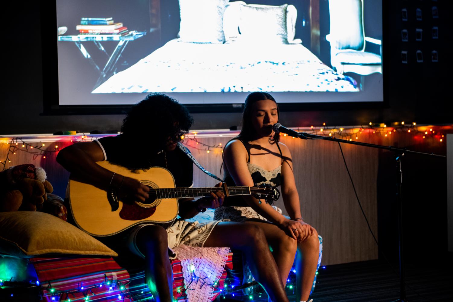 Two performers sit next to each other on a red futon. The one on the left is looking down while playing the guitar, while the other sings into a microphone.