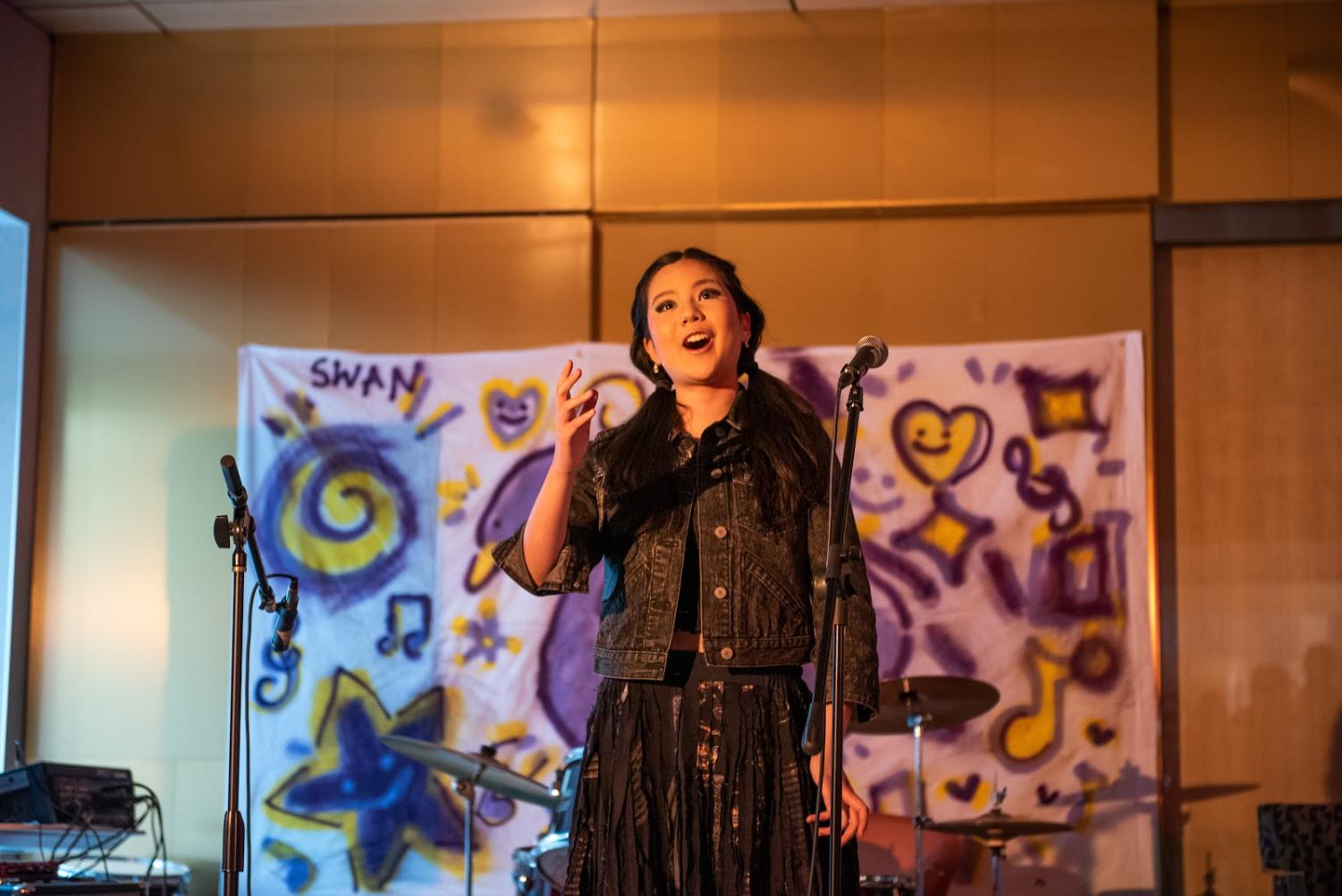 A singer with dark pigtails wearing a black denim jacket and a black batik skirt belts in front of a microphone.