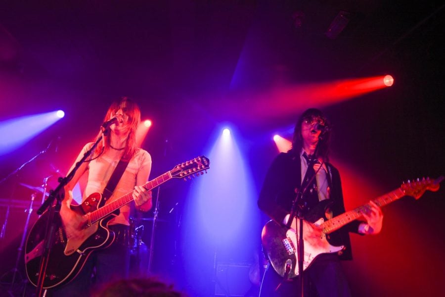 Two people with guitars sing into microphones next to each other, in front of red and blue stage lighting.