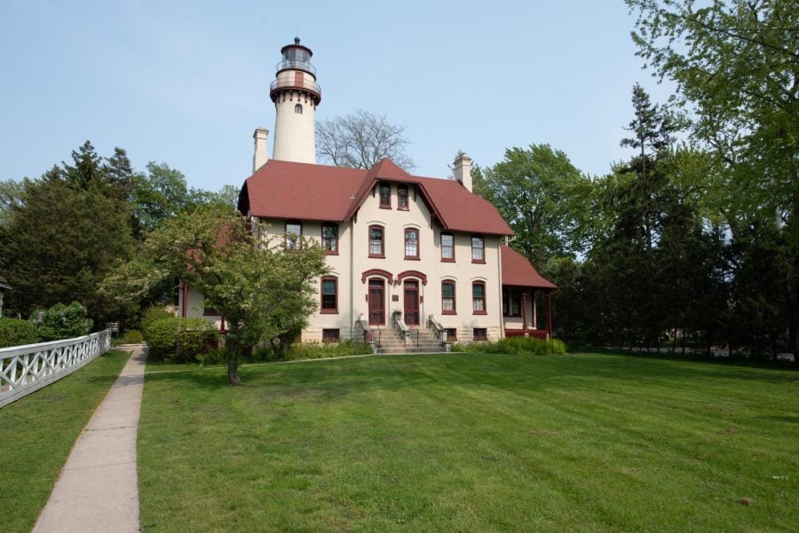 A white building with a red roof stands during the daytime in the center of the frame with a lighthouse behind it.