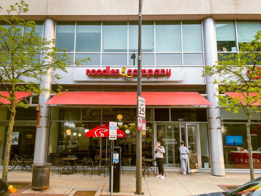 Exterior shot of Noodles & Company, with red lettering on the sign, a red awning covering a few dark brown tables and a red Noodles & Company branded umbrella. A person is standing in front of the restaurant, and another is opening the door to enter the restaurant.