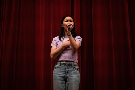 A person holding a microphone with their right hand over their heart sings in front of red curtains.