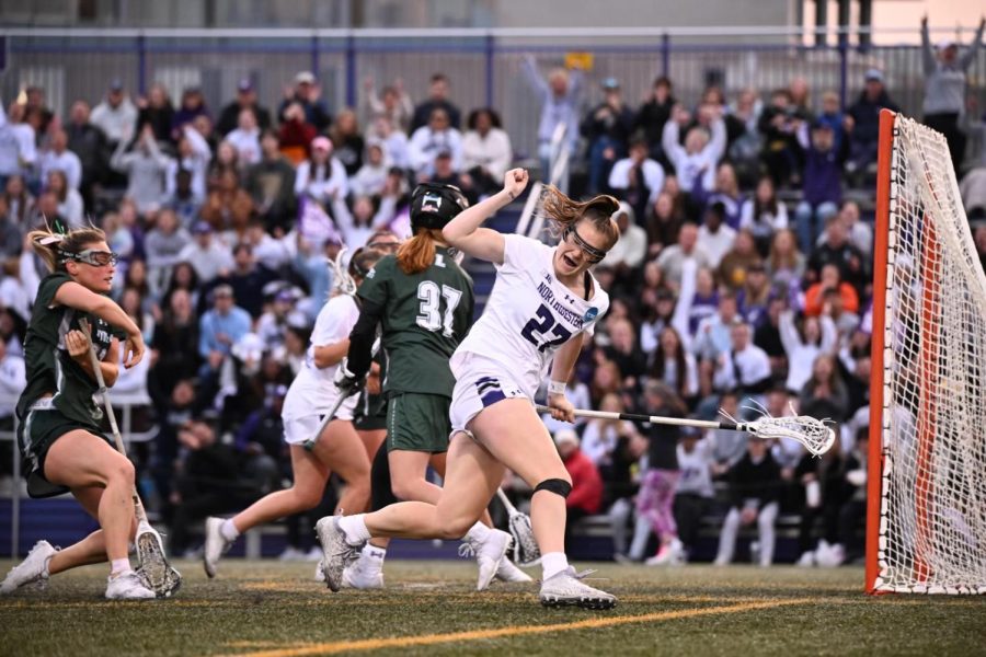 An athlete in a white jersey pumps her fist while holding a lacrosse stick.