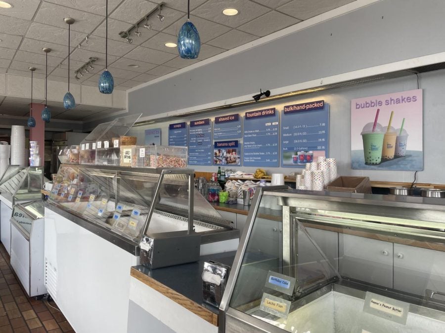 An ice cream shop. There are three serving stations with a cashier in between them. Above, blue lamps hang from a tiled ceiling. Behind are various blue signs displaying prices for menu items.