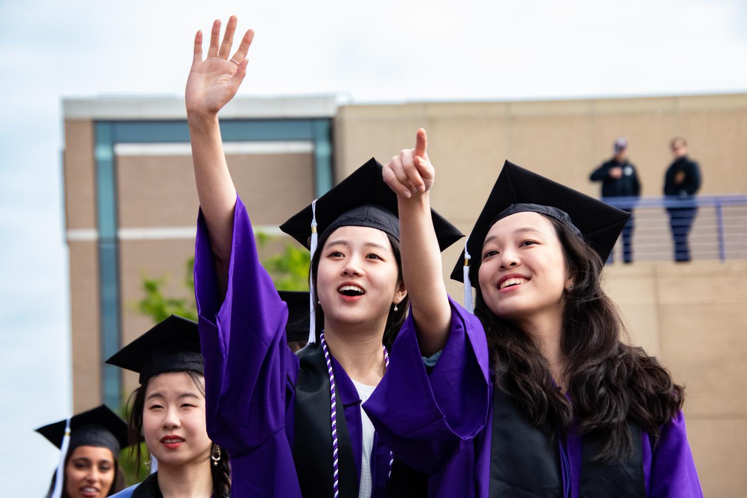 Two students in purple gowns and black caps point and wave to the crowd.