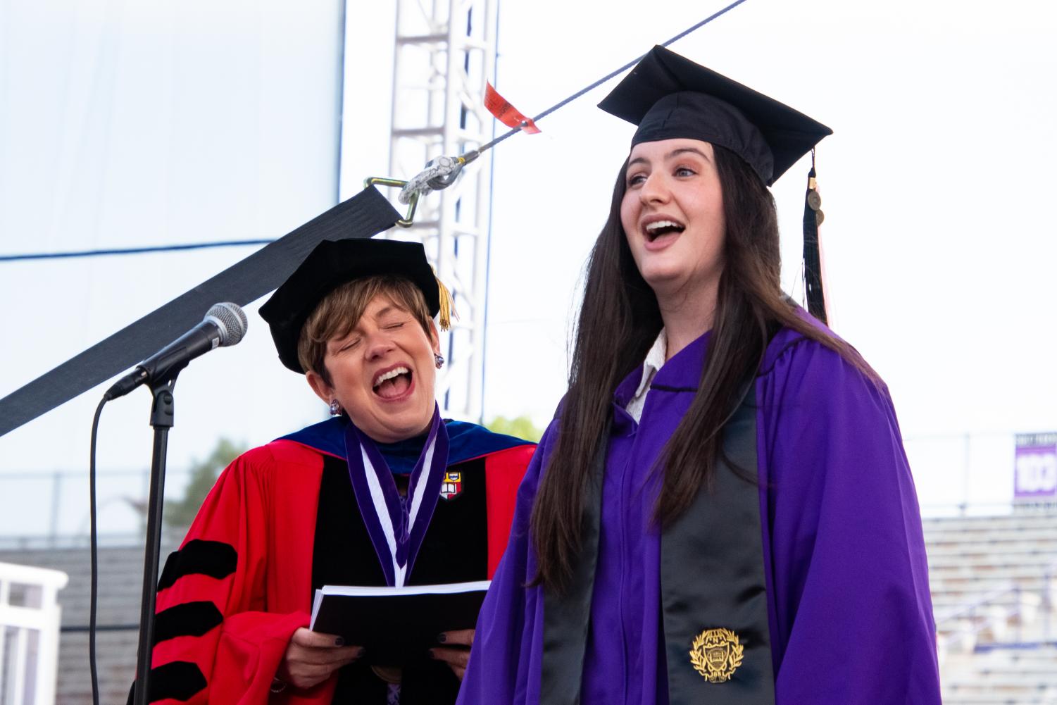 A student in a purple robe sings alongside another person in red.