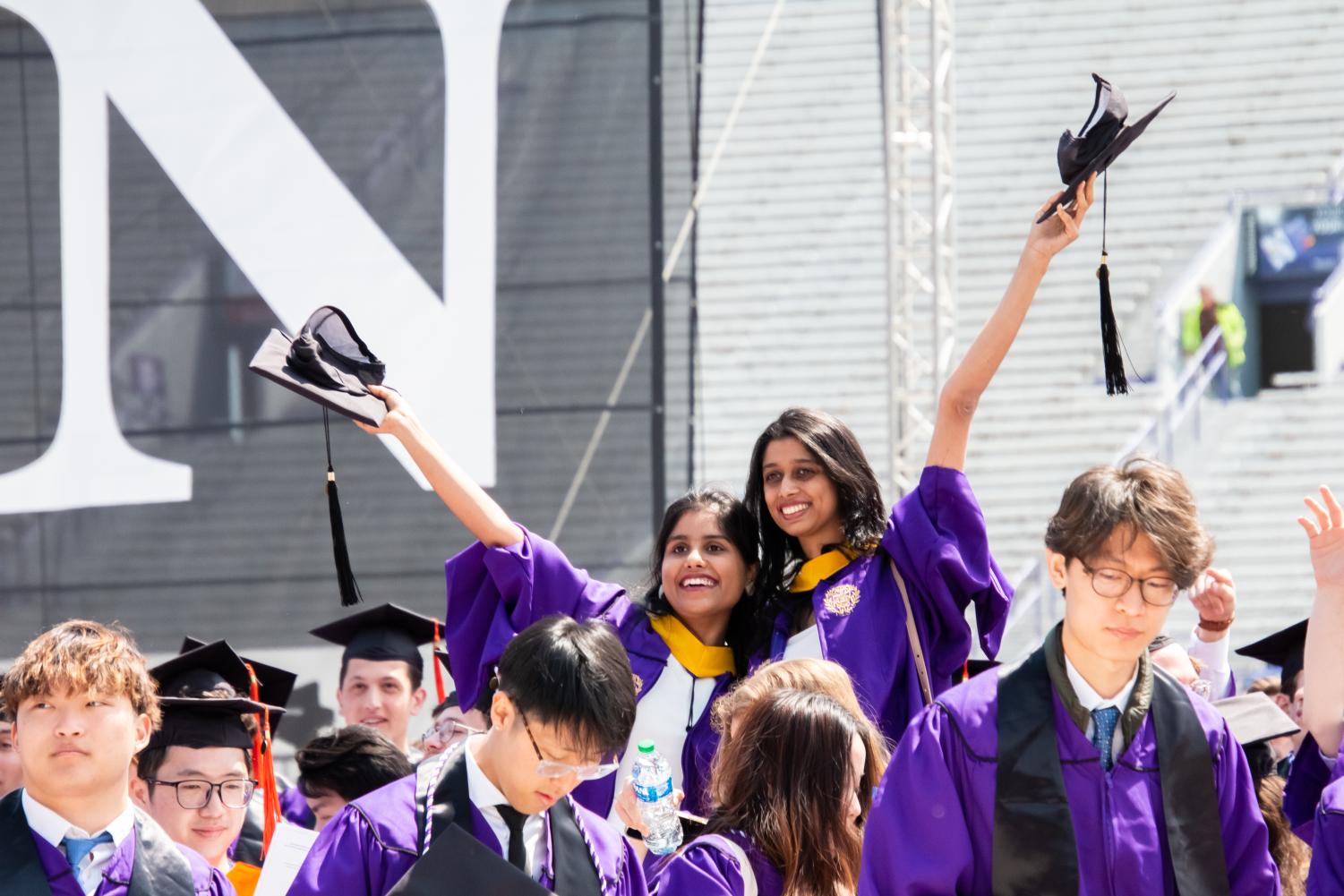 Two students with purple robes hold their arms up each student with a black cap in hand.