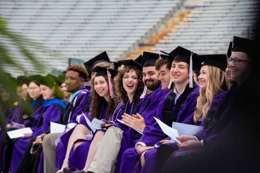 A group of 2023 graduates in purple robes smile and clap.