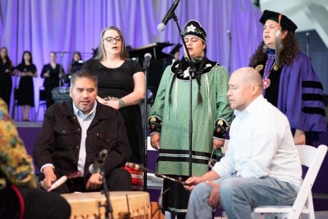 A group of five people sing around a drum. 