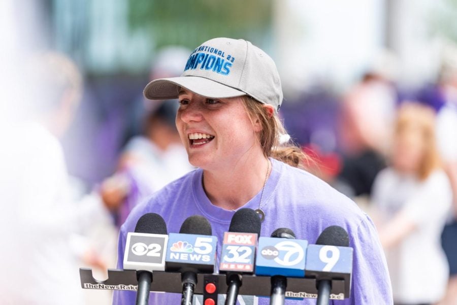 A woman in a purple shirt and gray hat speaks in front of microphones.