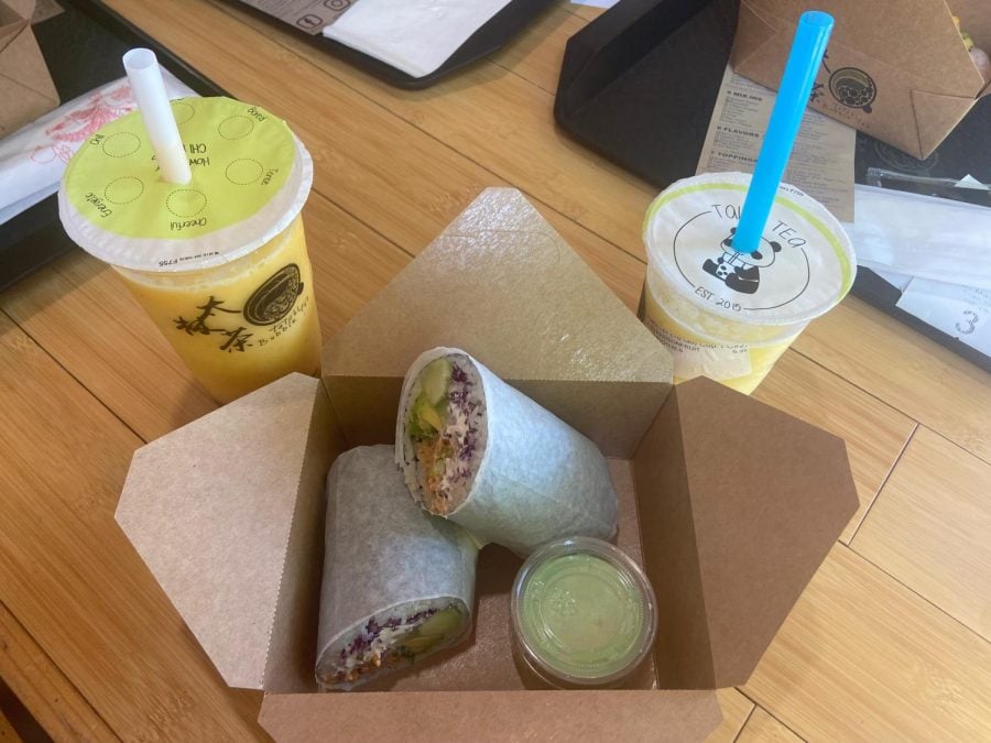 A sushi burrito cut in half in a brown box with two yellow fruit smoothies sitting behind it.
