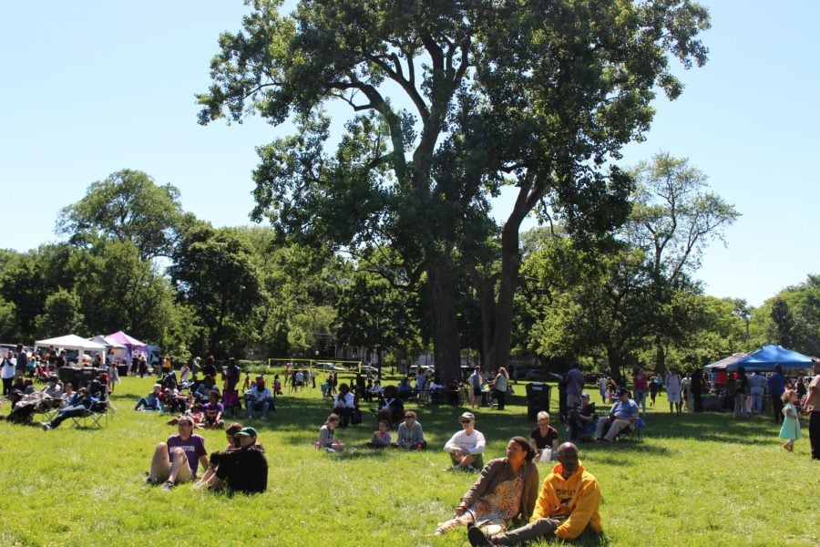 Groups of people sit on the grass facing forward, with tents and trees in the background.