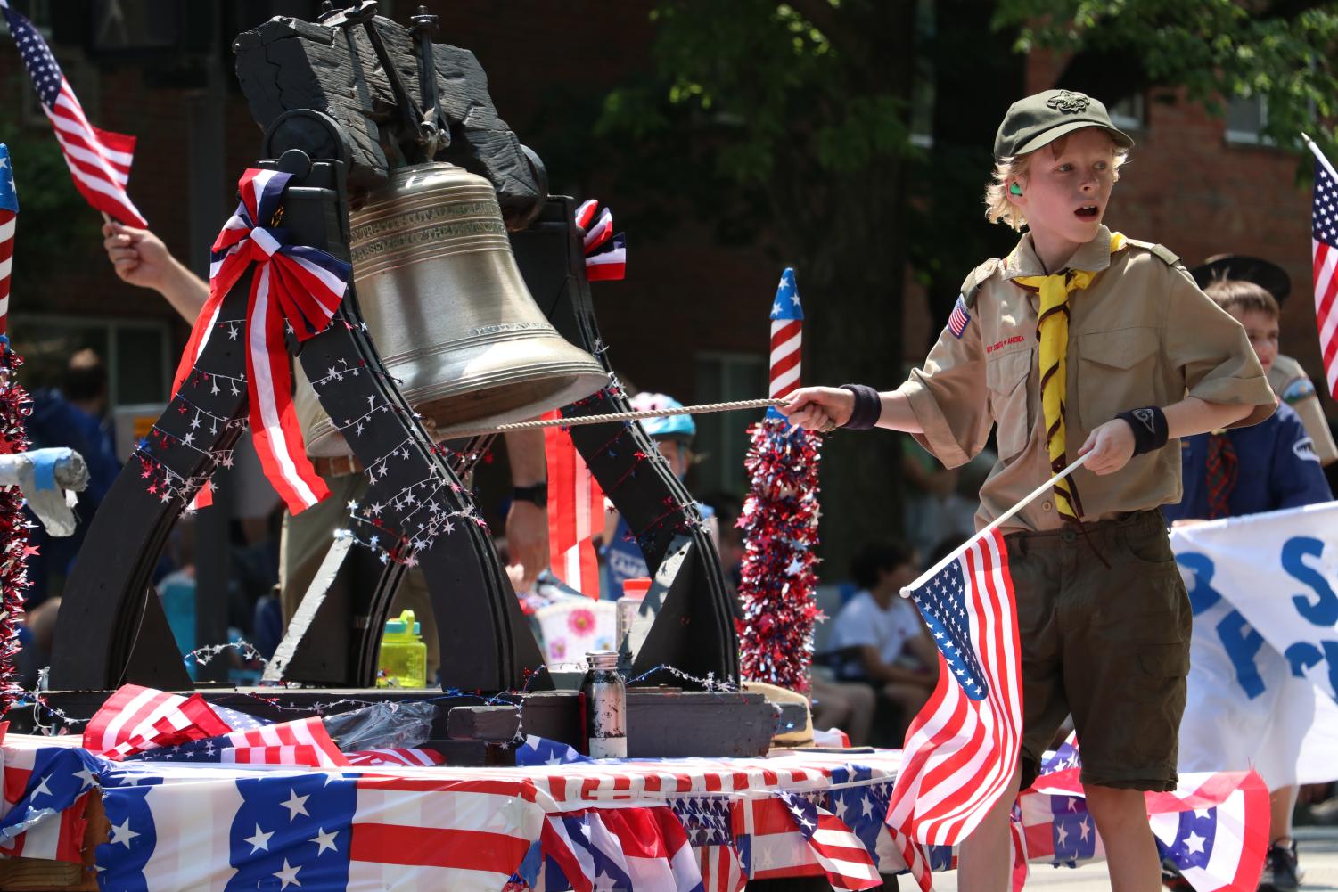 A boy scout rings a bell on a float decorated with American flags.