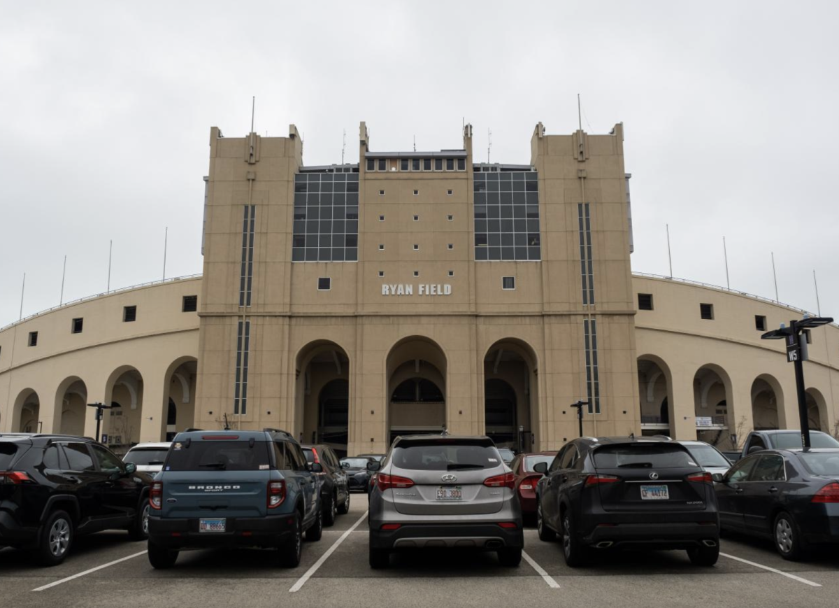 The exterior of Ryan Field, a large tan stadium with words reading Ryan Field and several cars parked in front.
