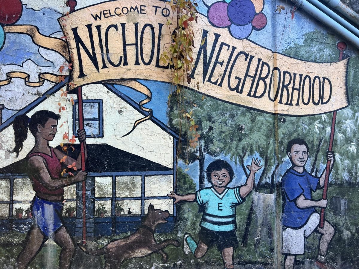 Colorful mural where two people hold a sign that says, “Welcome to Nichols Neighborhood.” A child in a soccer uniform and a dog dance under the banner.