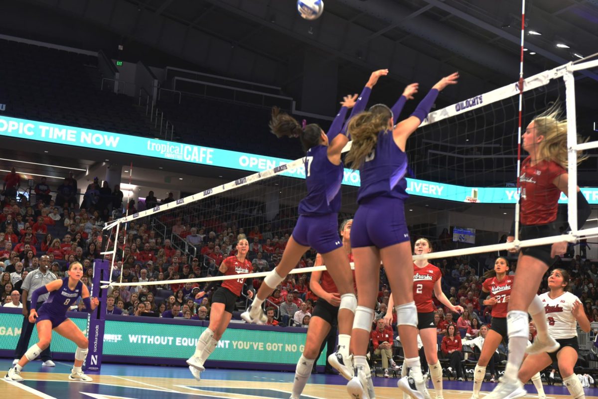 Two players in purple and white jerseys jump up to block a volleyball.