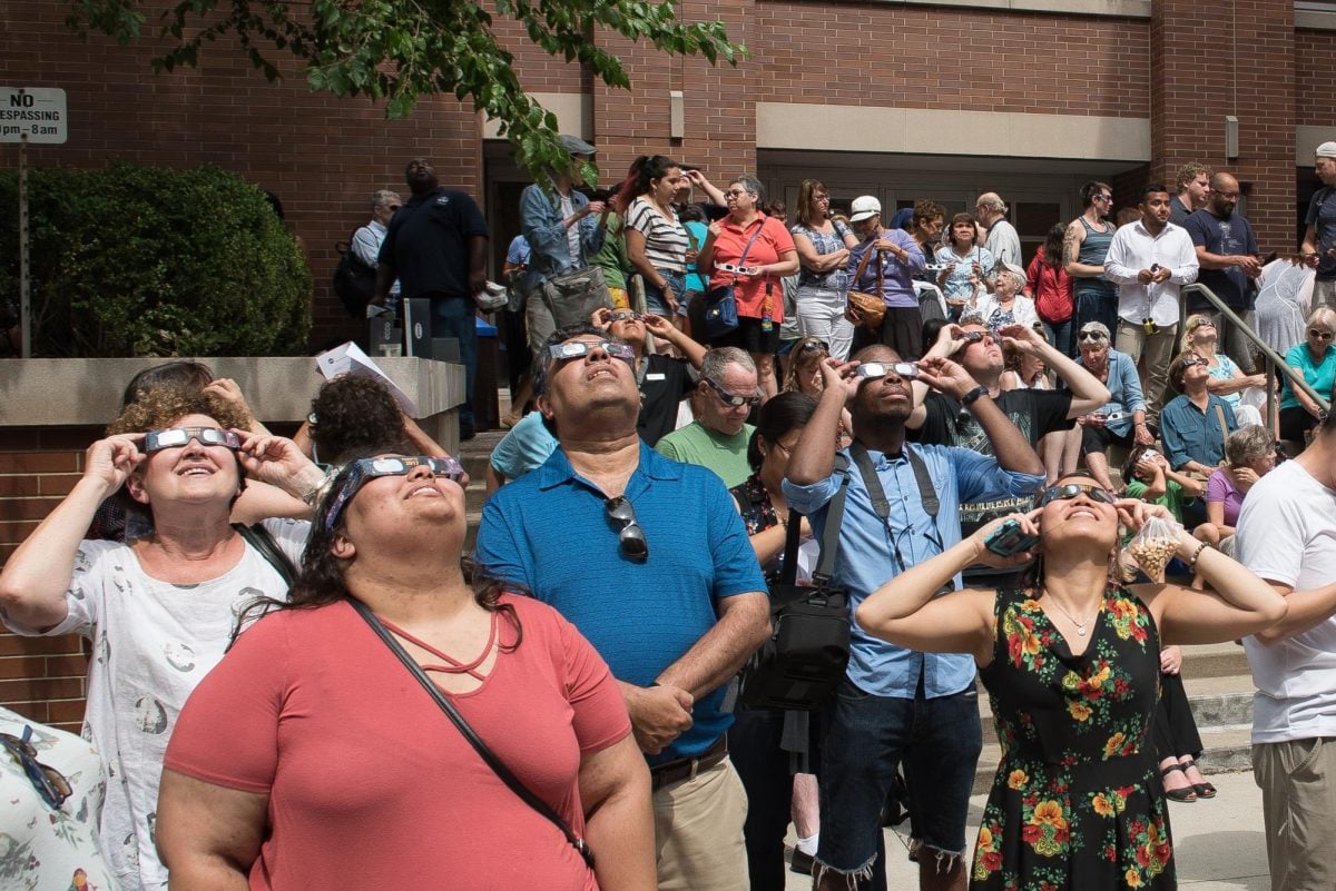 A group of people wearing eclipse glasses look toward the sky.