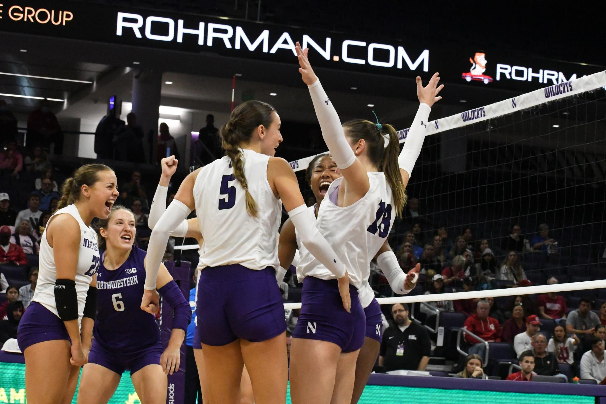 A group of players in white and purple volleyball uniforms celebrate.