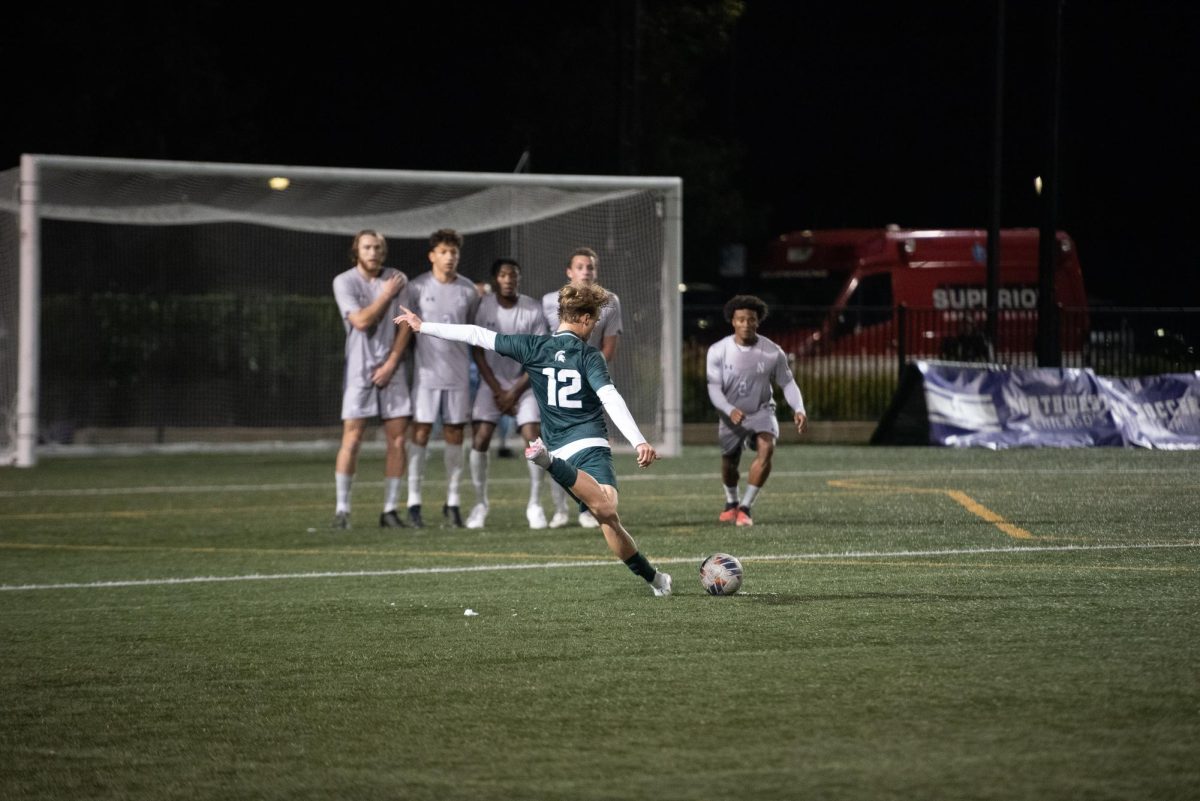 A player in a green jersey winds up to kick the ball around a wall of players in gray jerseys.