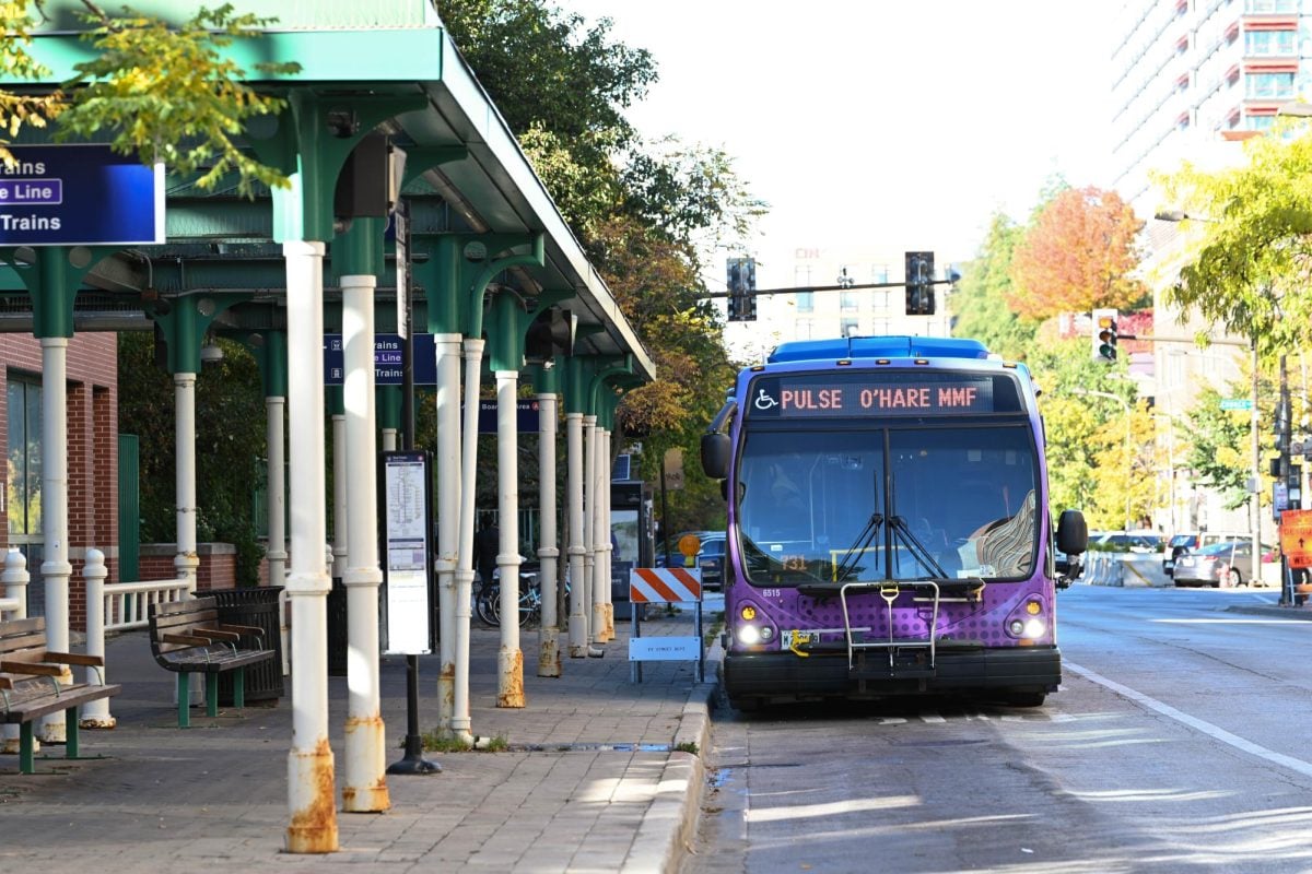 A purple bus sits next to a bus station with benches and awning.