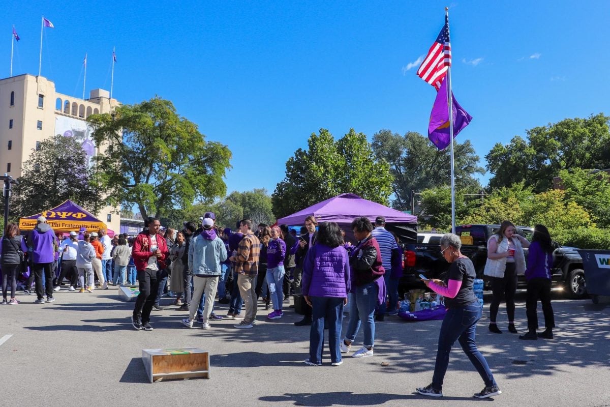 People+stand+around+purple+tents+and+a+game+of+cornhole+below+a+flagpole+with+two+flags.