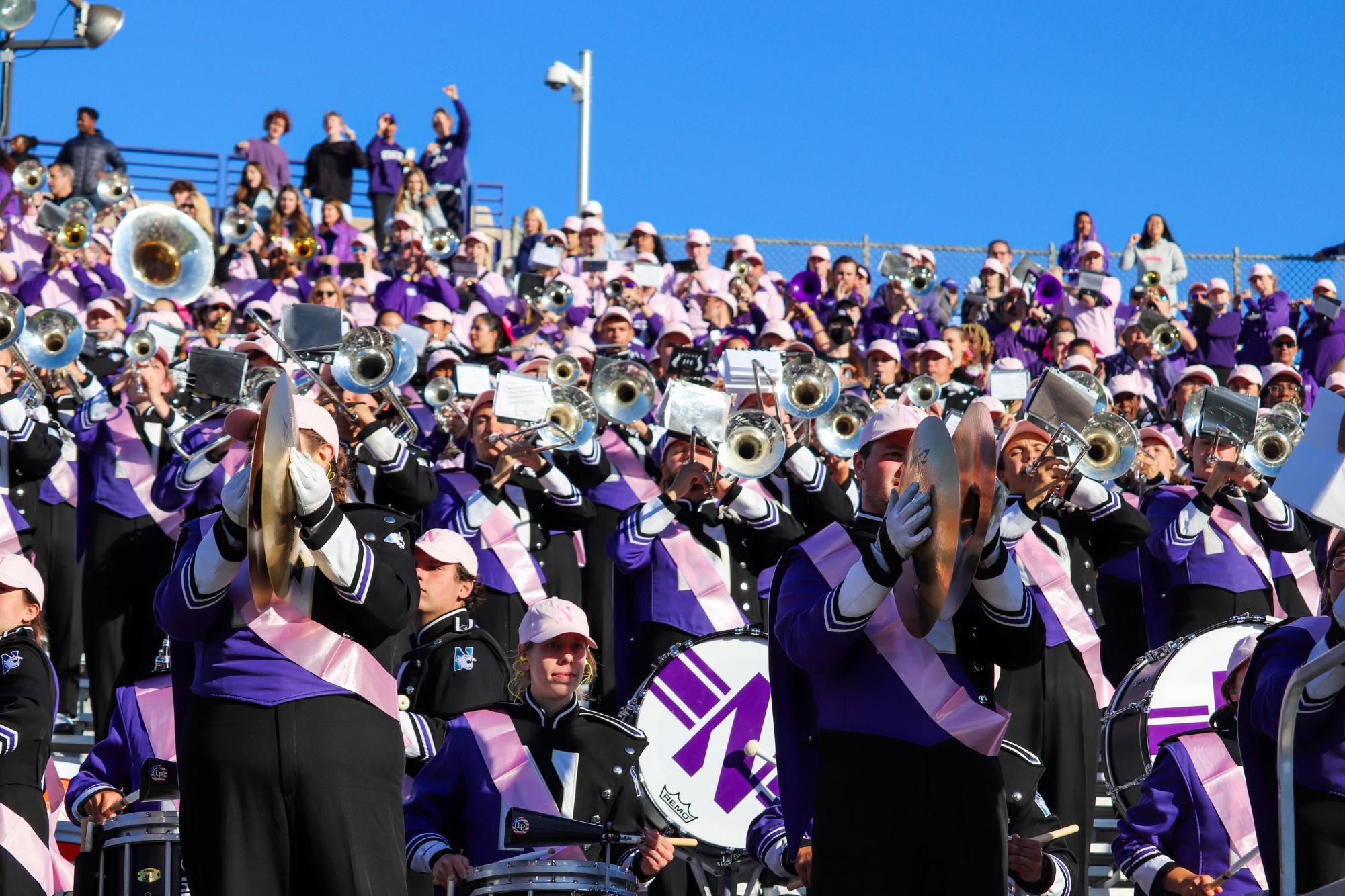 Marching band members in purple uniforms and pink sashes play their instruments.