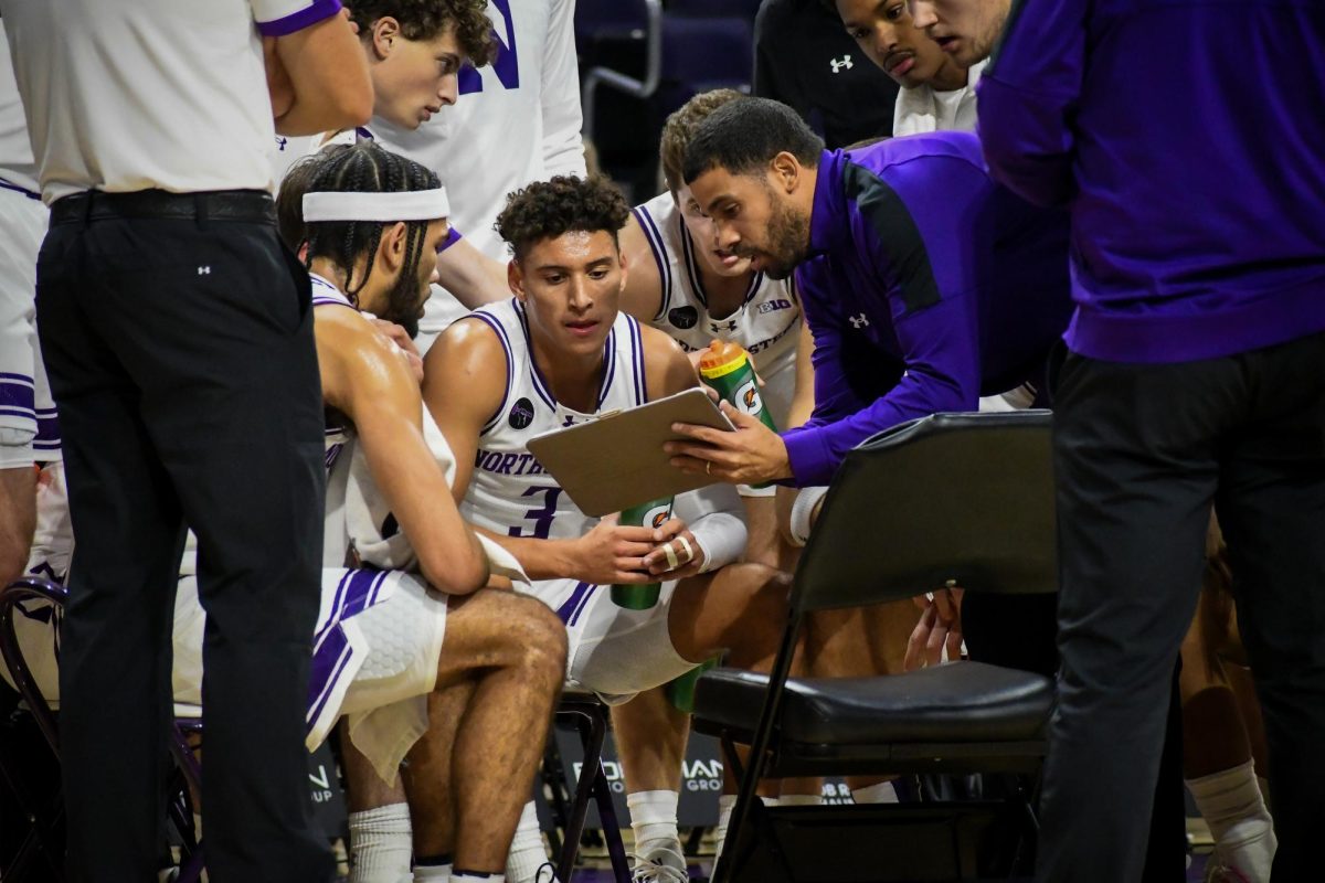 A group of players in white jerseys sit in a huddle next to a person with a clipboard.