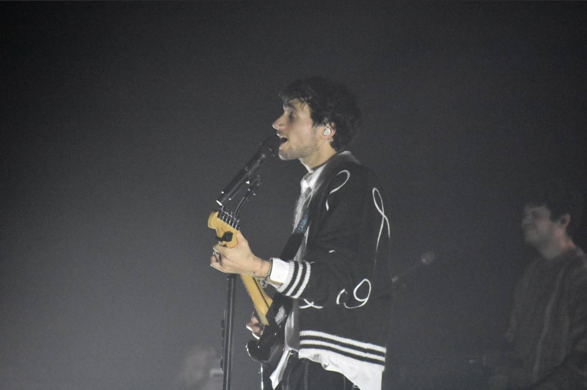 A person in black cardigan sings into a microphone while playing an electric guitar.

