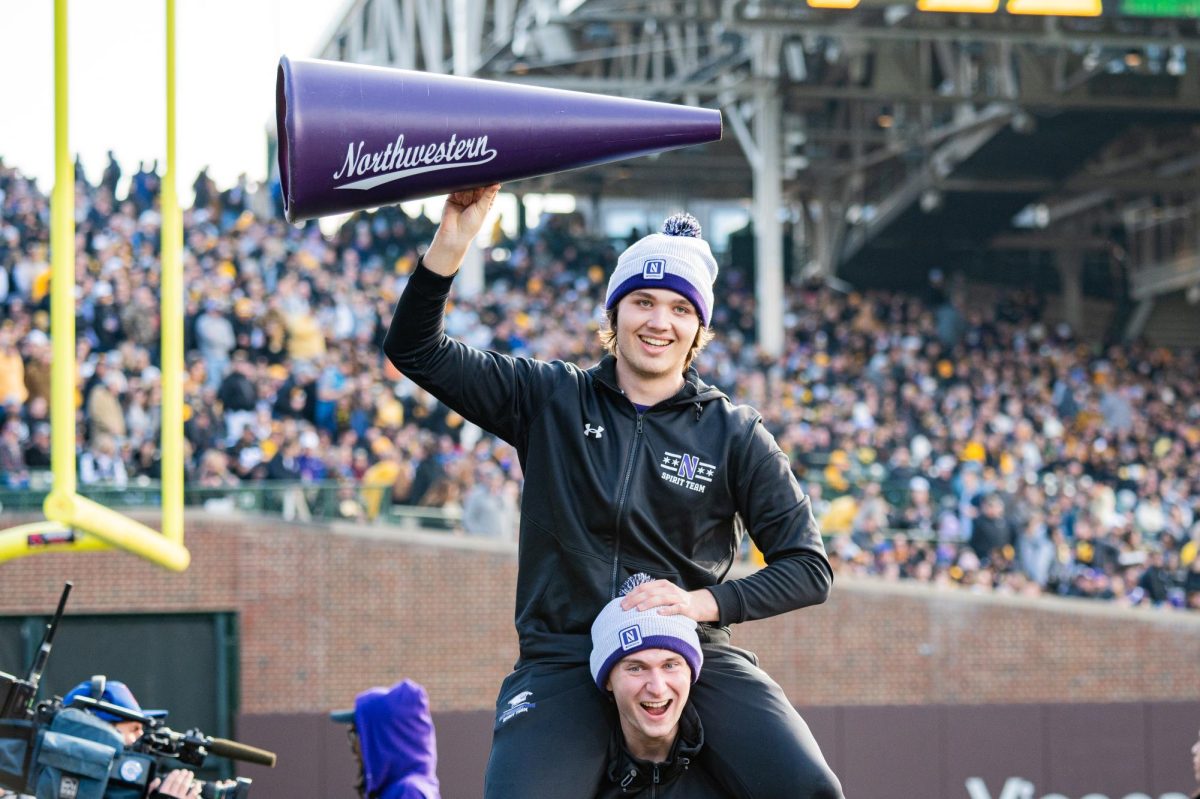 A person in black, sitting on another’s shoulders, holds up a purple megaphone.