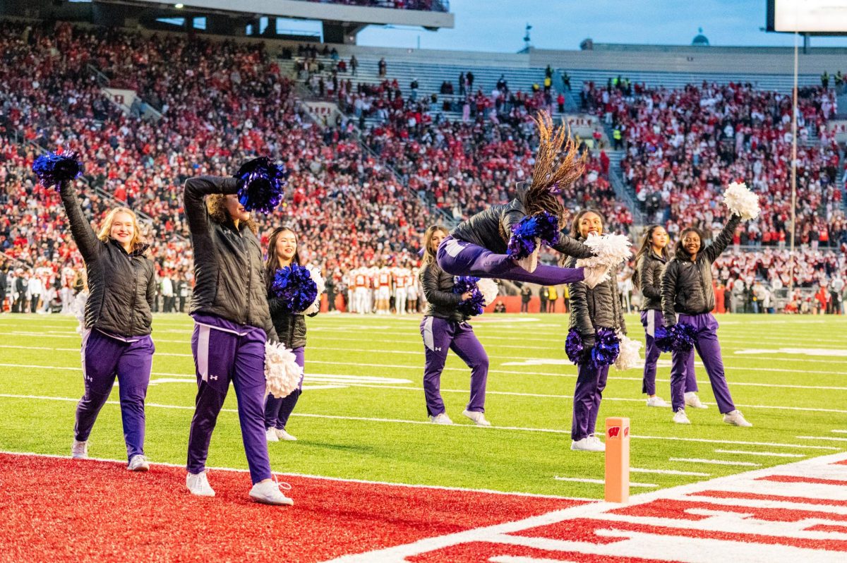 Cheerleaders in purple and black cheer on the field. One in the middle jumps in the air.