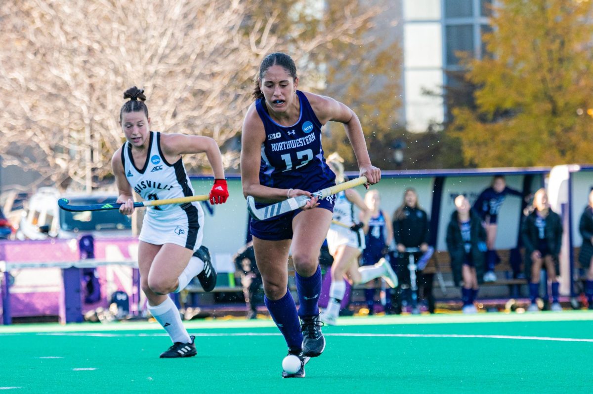 A field hockey player in purple and another player in white run after the ball.