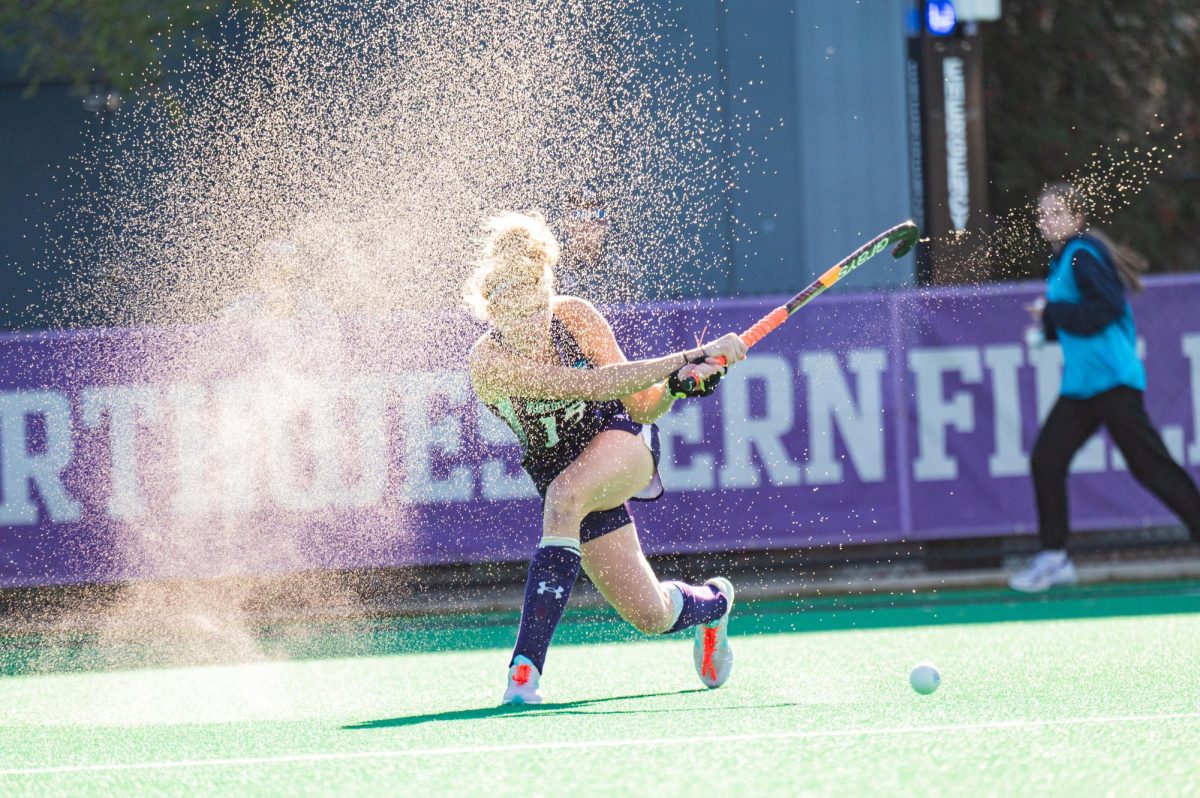 Water droplets sprinkle the air after a field hockey player in purple hits the ball.
