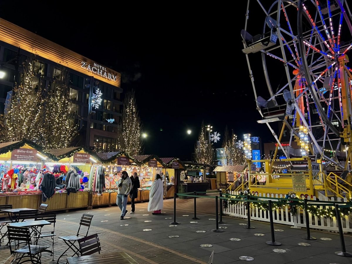 The+Christkindlmarket%E2%80%99s+wooden+vendor+stalls+at+nighttime+to+the+left+and+a+ferris+wheel+to+the+right.