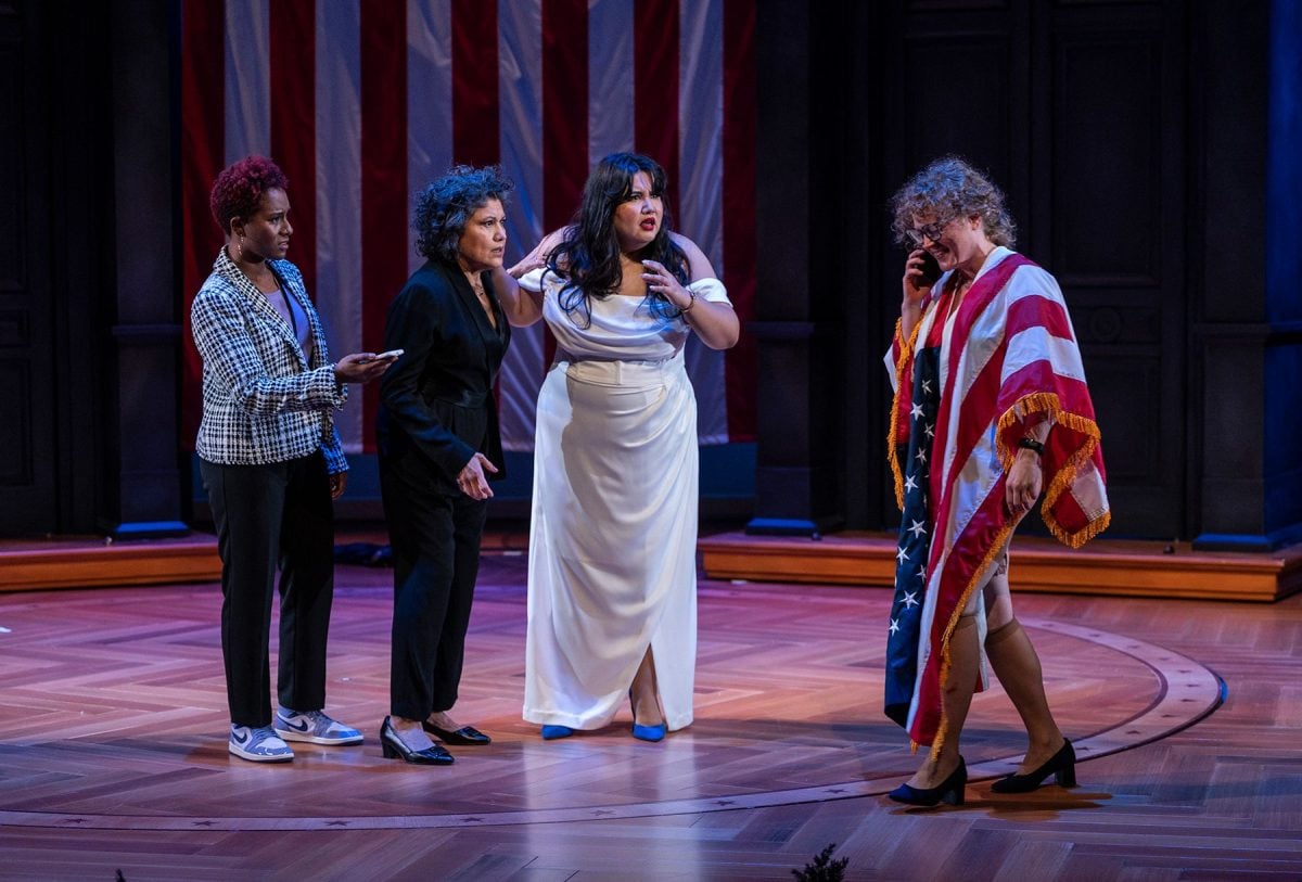 Three women confusedly watching a woman draped in an American flag as she talks on a cell phone.