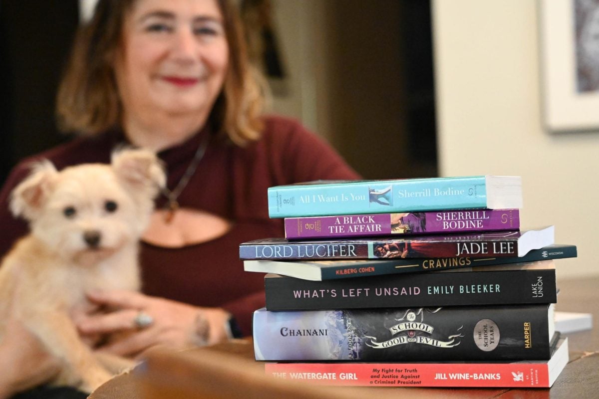 A stack of seven books sits atop a table where a woman sits with a dog in her hands.