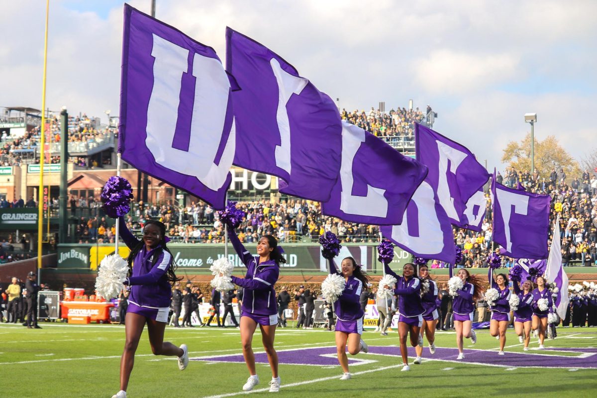 Cheerleaders in purple run on the field with purple flags that spell “Wildcat.”