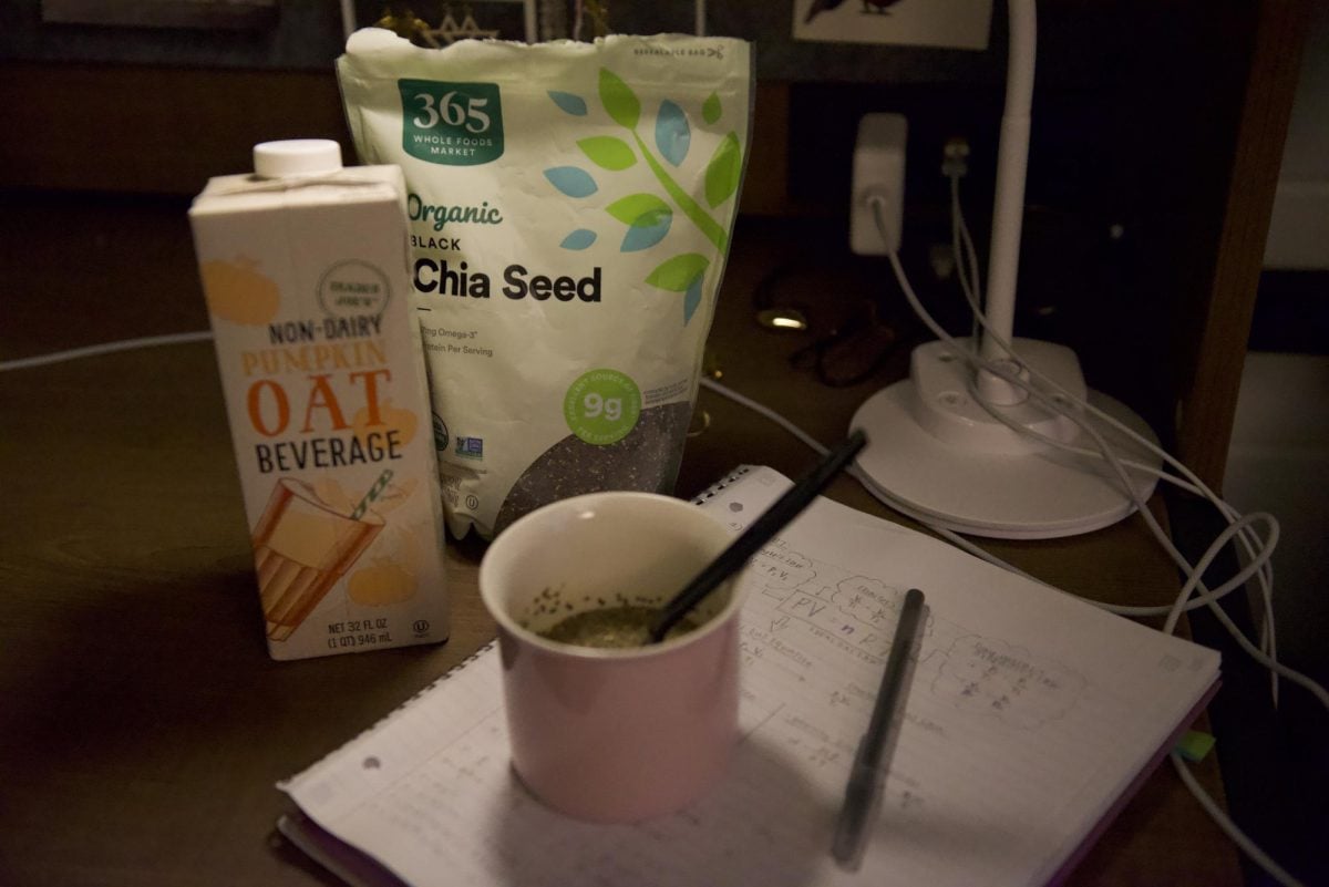 A mug with a spoon in it on a desk next to a carton of a pumpkin oat beverage, a bag of chia seeds and a notebook.
