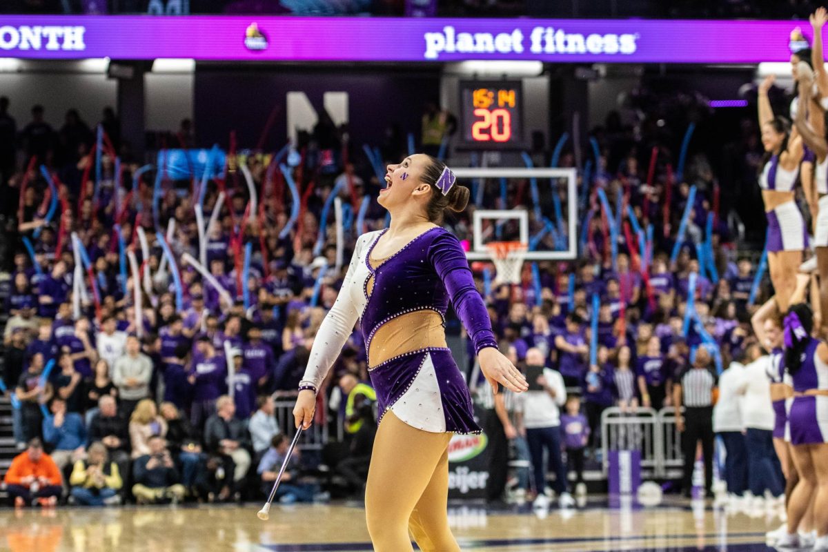 A student cheerleader in a purple and white outfit looks up and throws up a baton. 