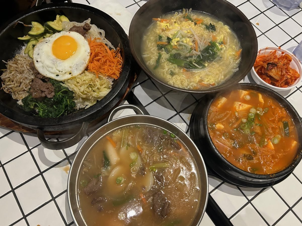 (In clockwise order) A pot of bibimbap with an egg on top, a bowl of veggie ramyun, a small white bowl of kimchi, a black pot of sundubu jjigae and a large silver pan of goong joong ddukbokki.