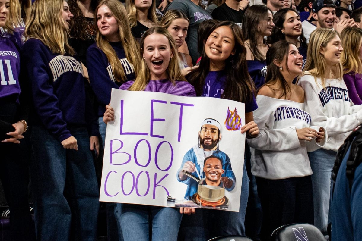 Two people holding a poster, surrounded by other fans. The poster says “Let Boo Cook” and features Buie’s head superimposed over a man cooking.