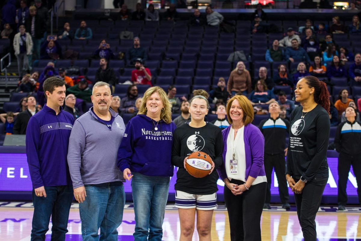 Six people pose for a photo in the middle of the court, all wearing Northwestern apparel.