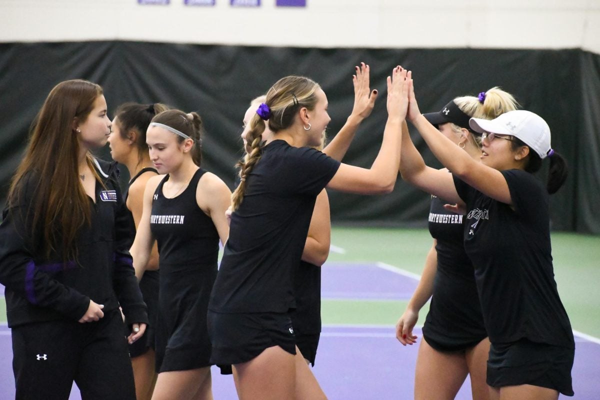 Two tennis players celebrate with a high-five, wearing black Northwestern attire. They are surrounded by other teammates.