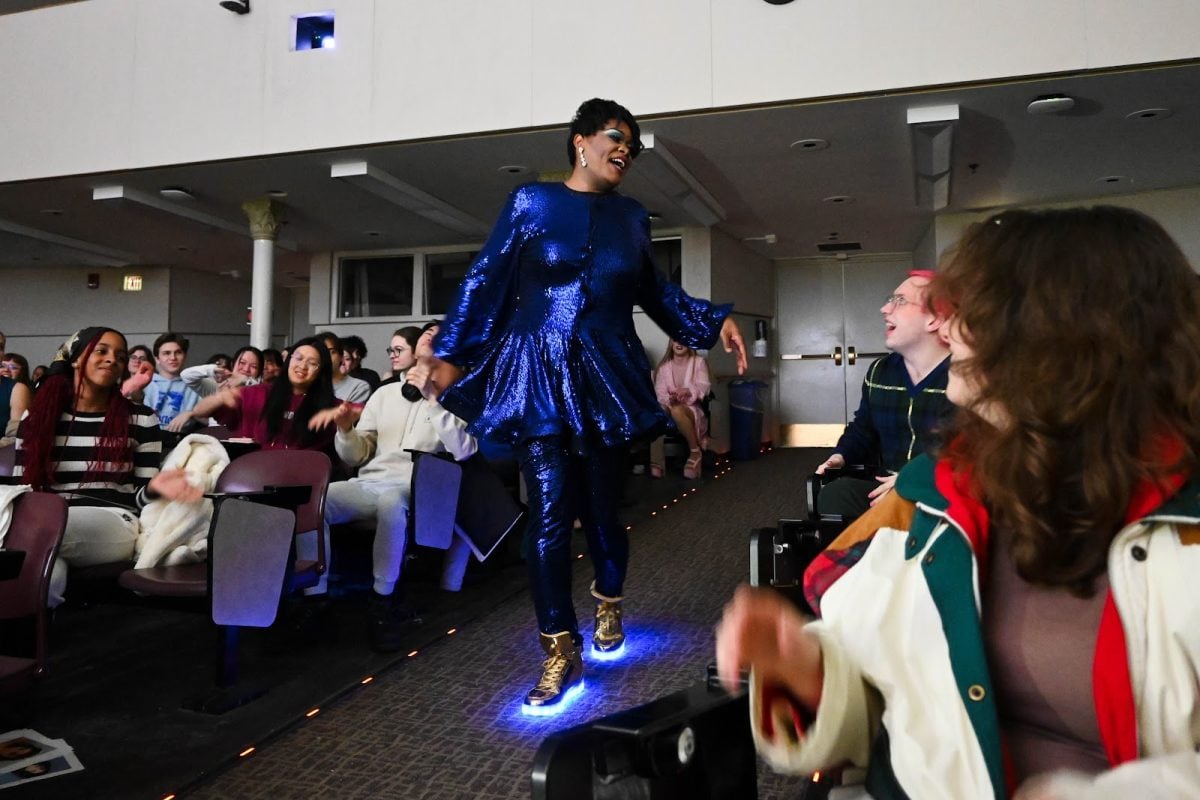 Drag queen Coco Sho-Nell, dressed in a blue sequined outfit, walks down an aisle with people on either side.