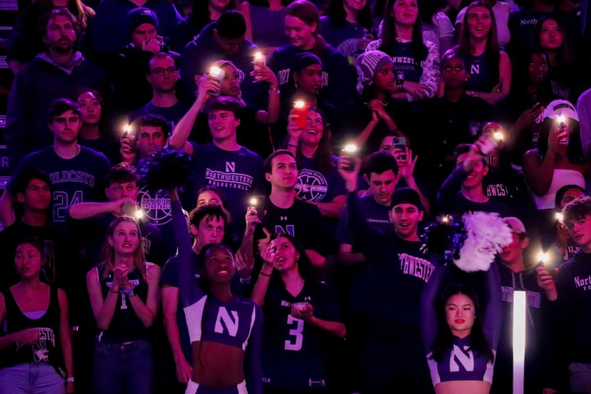 A crowd of people in bleachers stand in the dark and look forward. Some wave phone flashlights as purple light shines on them.
