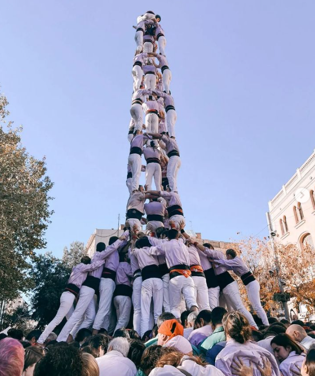 Many people wearing lavender shirts and white pants stand on top of each other, forming a human tower nine levels high.